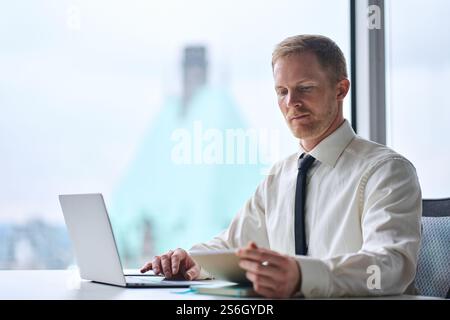 Young busy businessman manager working on laptop checking notebook in office. Stock Photo