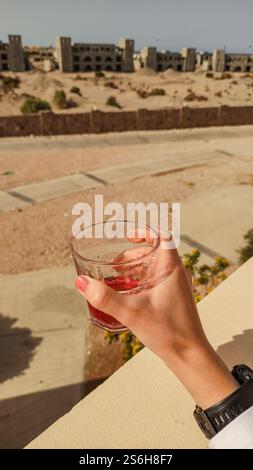 Hand with red nail polish holding a glass with red juice with a blurred view of an unfinished resort in marsa alam, egypt Stock Photo