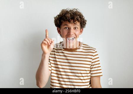 A young man with curly hair joyfully raises his finger in excitement, dressed in a striped t-shirt against a plain background Stock Photo
