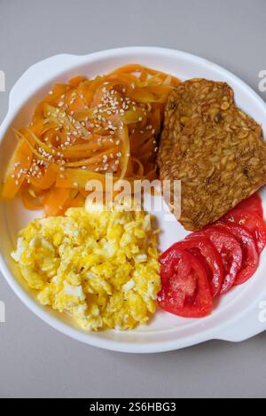 Easy carrot noodles, fried tempeh, tomato slices, and scrambled egg on a white plate - top view. Stock Photo