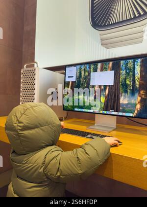 Paris, France - Dec 10, 2024: A child in a green winter jacket interacts with an Apple Mac Pro M4 silicon CPU and monitor in a retail store, with a se Stock Photo