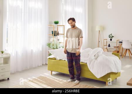 Young sleepy man suffering from sleepwalking standing with closed eyes near his bed at home. Stock Photo