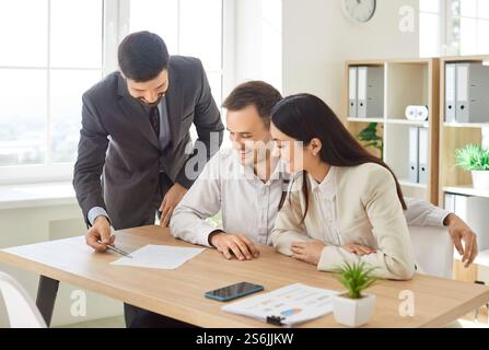 Confident young male realtor agent pointing at paper for family couple of customers, clients signing Stock Photo