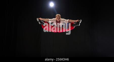 ale dancer in bright red trousers performs airborne split jump, arms outstretched, showcasing explosive athleticism of Hopak, against black studio Stock Photo