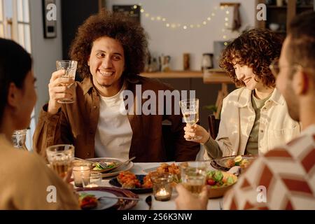 Curly haired young adult biracial man holding glass with apple juice while smiling widely during funny and warm conversation with his friends Stock Photo