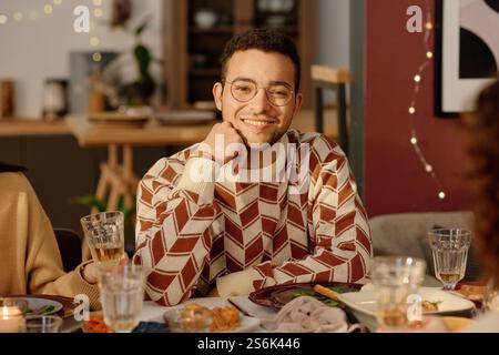Medium close up portrait of young adult biracial man dressed in sweater with ornaments leaning his head on hand and smiling while looking at camera Stock Photo