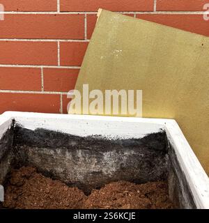 A white container filled with soil, placed against a brick wall with a golden board leaning, representing gardening or construction concepts. Stock Photo