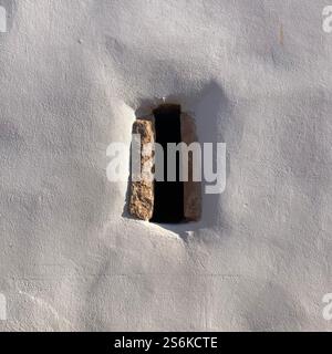 Detail of a narrow window with a stone frame against a textured white plaster wall, showcasing timeless rustic design and natural lighting contrast Stock Photo