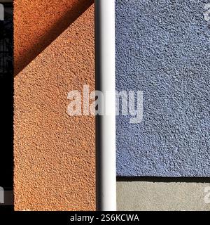 Vivid contrast of orange and blue textured wall surfaces divided by a vertical white pipe in daylight, creating a minimalist geometric ambience. Stock Photo