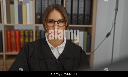 Woman judge in courtroom wearing glasses and black robe, sitting in front of bookshelf, concentrating on her work with a serious expression Stock Photo