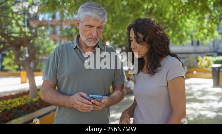Middle-aged couple standing outdoors in an urban park, man showing smartphone to woman, both appearing engaged in conversation against a backdrop of t Stock Photo