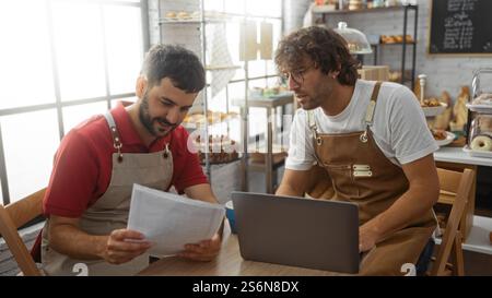 Men working together at a bakery shop, looking at documents and a laptop, surrounded by shelves of bread and pastries, engaging in conversation and pl Stock Photo