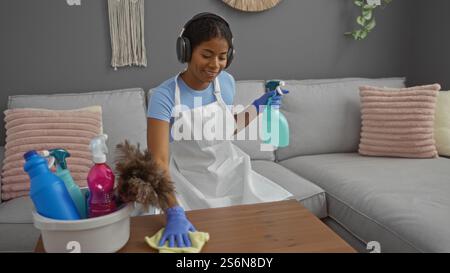 Young woman cleaning living room with cleaning supplies while wearing headphones indoors at home Stock Photo