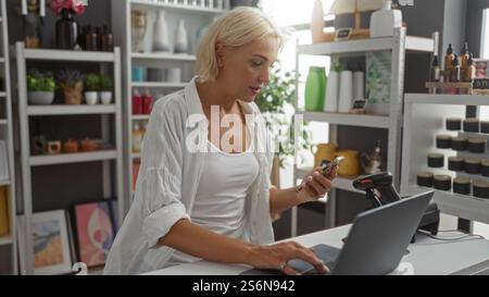 Blonde woman holding a phone and working on a laptop in a beautifully decorated home decor store with shelves of various items in the background Stock Photo