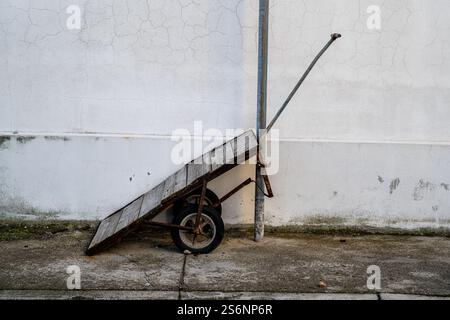 An old, rusty hand truck leans against a weathered, white stucco wall. No one is visible in the image, but the presence of the cart suggests it was li Stock Photo