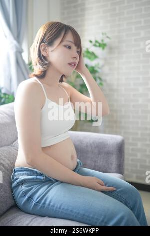 A pregnant Japanese woman in her 20s wearing clothes that show her stomach is sitting on a sofa in her apartment, holding her big belly. Stock Photo