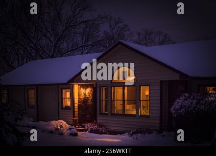 View of Midwestern suburban house with large windows after snowfall at night; windows lit and snow on ground, roof and trees Stock Photo