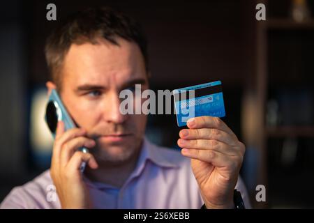 Businessman business man talking on phone during call hold card. A young man in a pink shirt is dictating CVC code from a card to a phone fraudster. b Stock Photo