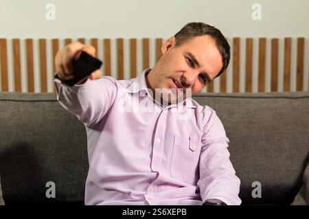Pleased unshaven man changes TV channels with remote control watches funny program dressed in casual clothes sits crosses legs on comfortable sofa iso Stock Photo