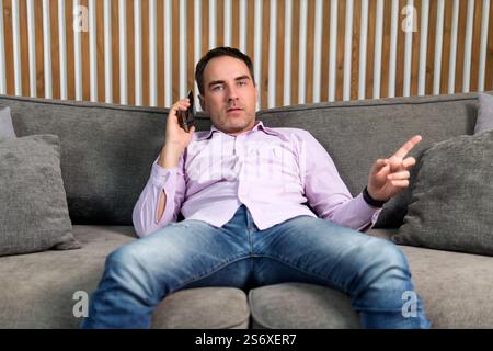 Home-office businessman talking on phone. A young man is talking on the phone and gesturing with his hands while sitting on the couch. Stock Photo