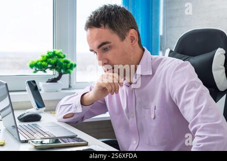 A handsome young man in a pink shirt is sitting at a desk in a bright office and working hard at a computer. An employee reads messages on his smartph Stock Photo