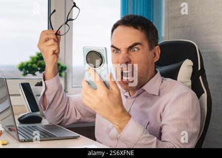 Angry male arguing and screaming on mobile phone while working on laptop online at home. Annoyed man freelancer shouting into the phone gesturing, des Stock Photo