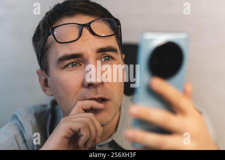 A young, handsome office employee looks at the screen of his smartphone with amazement, surprise and interest and carefully reads incoming messages. Stock Photo