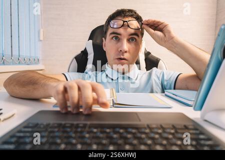A young handsome man in a blue shirt is working at a computer on a light background. the nerd looks at the monitor screen Stock Photo