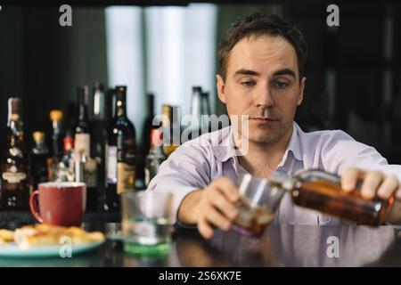 A young man drinks whiskey from a glass in a bar on a dark background. A businessman with a bottle in his hands is relaxing in a pub with alcohol afte Stock Photo