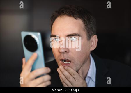 Portrait of surprised young man reading message on smartphone, look curious at mobile screen, standing against dark background Stock Photo