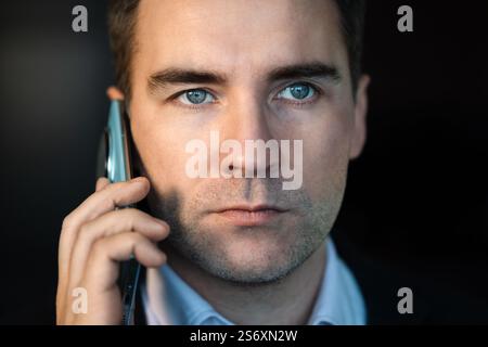 Young Businessman Talking On Phone At Night In Modern Office. close-up of a young man's face talking on the phone against a dark background at night. Stock Photo