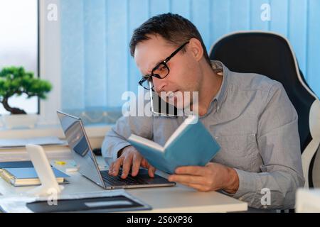 Focused man in glasses and blue shirt working at home with laptop, writing in notebook, doing remote work or study sitting in bright office Stock Photo