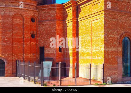Red brick building with intricate architectural details, arched windows and fence in foreground. Structure is illuminated by sunlight highlighting its Stock Photo