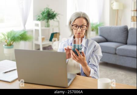 Senior woman working at the desk with laptop holding mobile phone in hands. Stock Photo