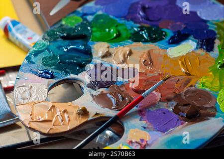 Artists palette with mixed colorful paints, brushes, and paint tubes in the background. Stock Photo