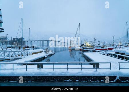 Snowy harbor docks with moored boats and yachts overlooking the distant Tromso Bridge under a cloudy winter sky in Tromso, Norway Stock Photo