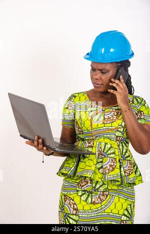 beautiful young female engineer wearing hard hat holding laptop and talking on mobile phone. Stock Photo
