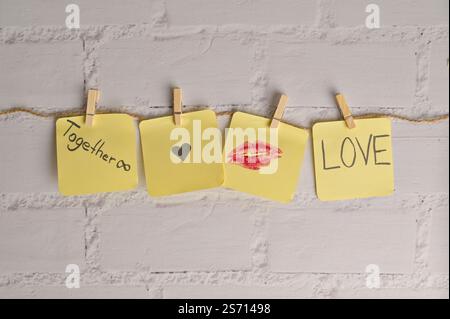 Four yellow sticker papers with lipstick kiss mark, inscription "Love", "Together" and small black heart on a string with clothespins, white brick Stock Photo