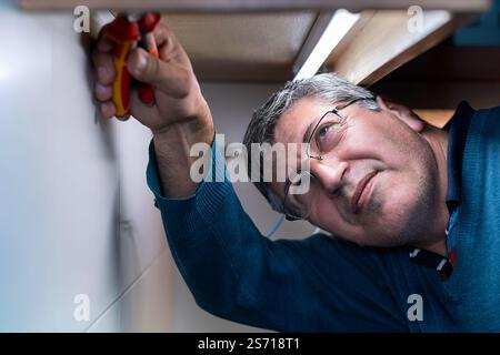 Man focused on repairing electrical fixtures under a cabinet using pliers, performing precise maintenance work. Stock Photo