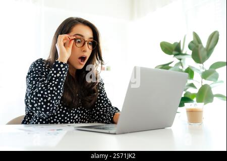 Surprised business woman with glasses looking at a laptop screen with amazement, sitting at a desk with documents and coffee in the office while working on a project, showing astonishment Stock Photo