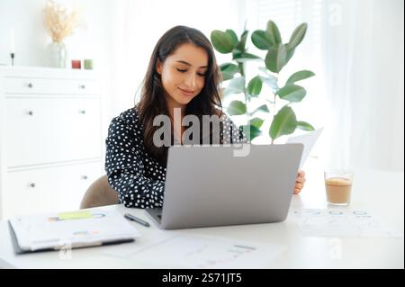 Confident beautiful caucasian or arabian brunette woman, working on a laptop while holding a document, sitting at a desk with charts and a cup of coffee in a bright modern office, showing focus and Stock Photo