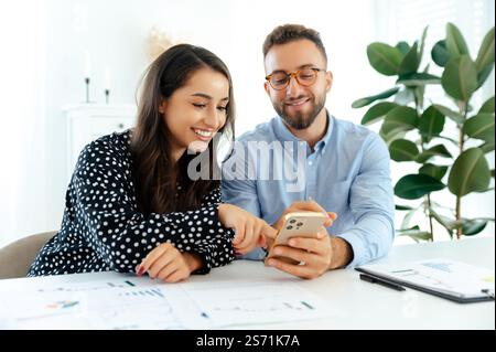 Happy multiracial business colleagues sitting at a desk with charts and a laptop, pointing at a smartphone together in a bright modern office, sharing ideas, discussion future strategy, smiling Stock Photo