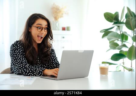Excited arabian or caucasian business woman smiling happily while looking at a laptop, sitting at a desk with charts and a coffee cup in the office, showing amazement and joy in a modern bright office Stock Photo