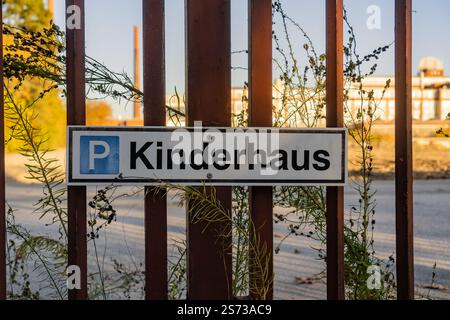 A sign with the word Kinderhaus on it is hanging from a fence. The sign is surrounded by weeds and is located in a parking lot Stock Photo