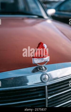 Close-up of a Mercedes-Benz W123 hood ornament wearing a small red Santa hat, set against the classic orange car in an outdoor parking area in Prague. Stock Photo