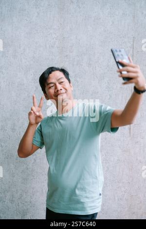 Asian man taking a selfie with a peace sign against a gray background. Stock Photo