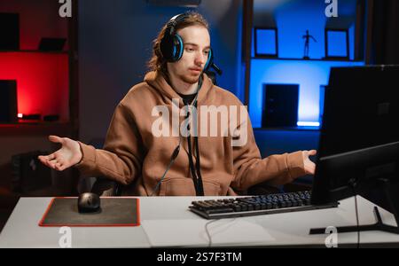 Confused Young Caucasian hipster pro gamer shoulders up say can't help, makes gesture of I dont know, playing in online cyber sport game at night at h Stock Photo