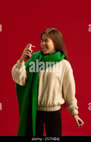 Teen girl in green scarf and beige sweater suffering from cold and flu, using throat spray to reduce soreness isolated on red background. Stock Photo