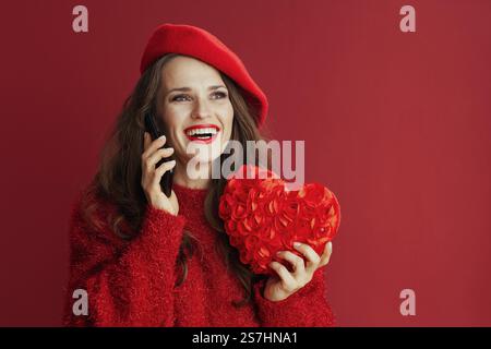 Happy Valentine. happy modern 40 years old woman in red sweater and beret with red heart speaking on a smartphone. Stock Photo