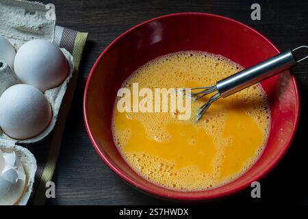Whisking eggs. Whipped raw eggs in a red bowl with whisk for omelet , close up. Breakfast preparation, rustic style Stock Photo